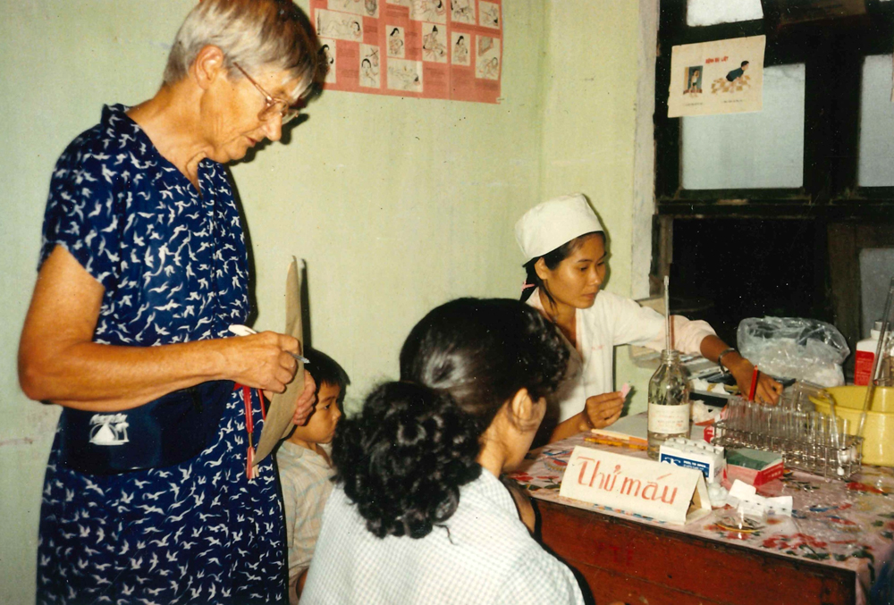 Woman in blue dress and glasses in conversation with two Asian women wearing white medical uniforms.