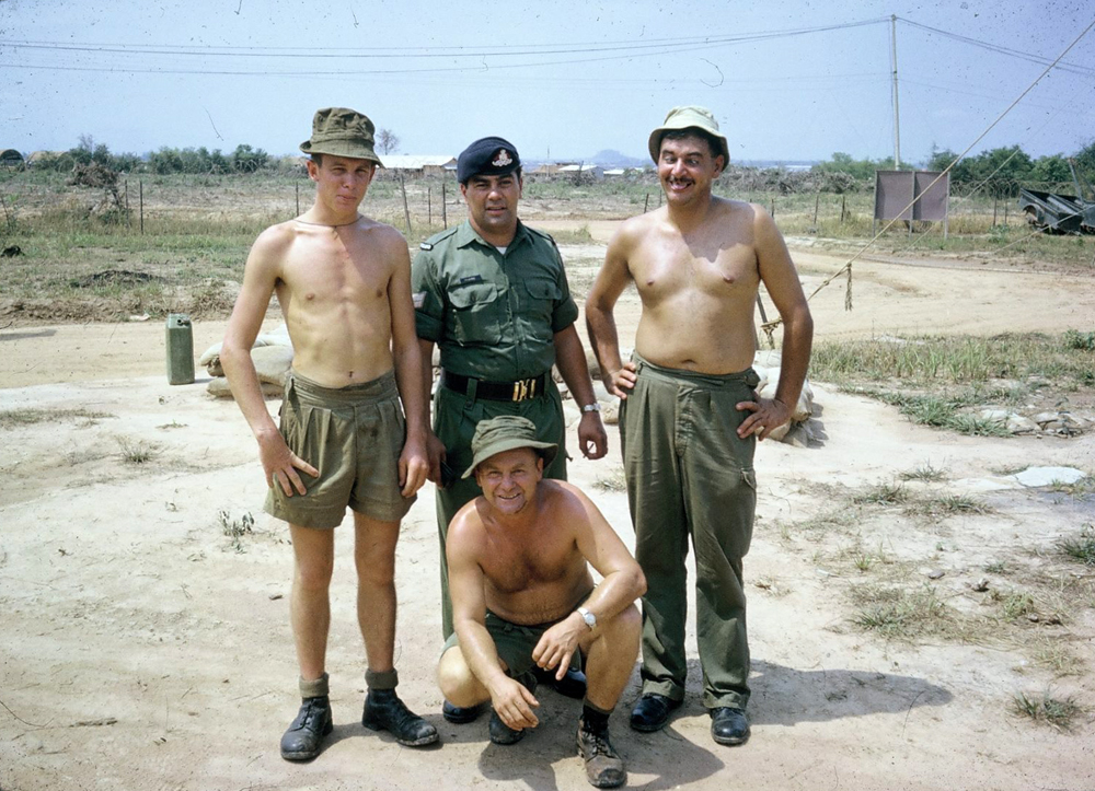 Four men, three shirtless, grouped together in military uniform.