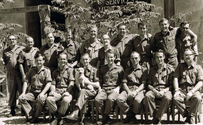 Black and white photograph of men in uniform sitting in two rows. One is holding a white dog.