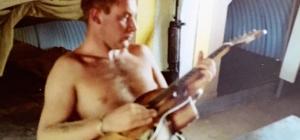 Bare chested man sitting at table holding a guitar.
